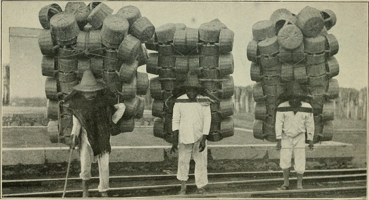 Three men carrying enormous stacks of woven baskets on their backs beside railway tracks